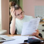 Indoor shot of young European Caucasian girl looking at financial documents at home with deeply bored face looking sick and tired of her economic problems, trying to check counts and all details