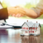 Real estate agent and customer sign contract papers and shake hands with small model home in front.