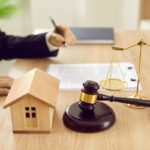 Attorney sitting at desk with scales of justice, gavel and small wooden toy house, working with documents, signing contract agreements. Real estate law, foreclosed property, lawyer services concept
