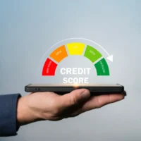 A hand holds a smartphone projecting a credit score gauge from very poor (red) to excellent (green) with "CREDIT SCORE" text. Background is light blue. A hand holds a smartphone projecting a credit score gauge from very poor (red) to excellent (green) with "CREDIT SCORE" text. Background is light blue.